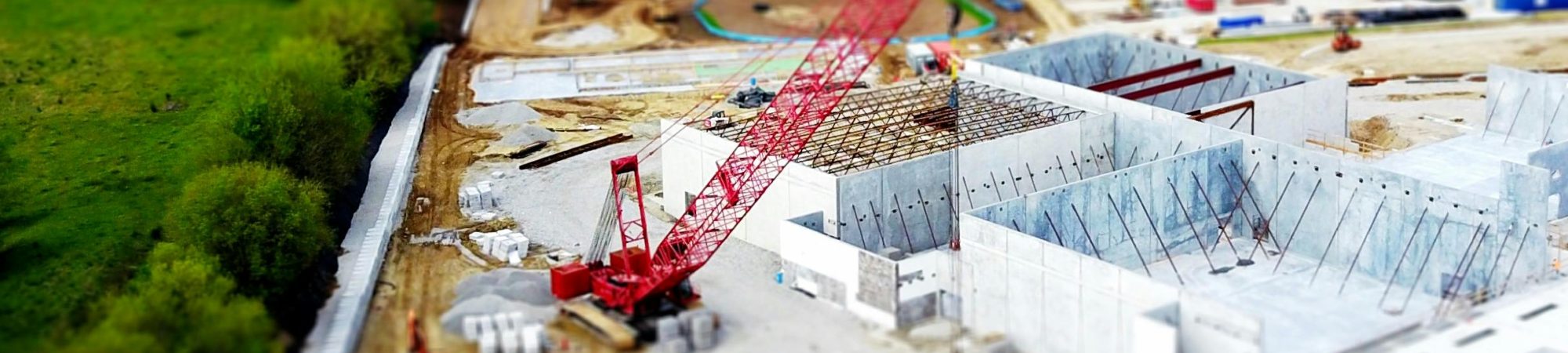 Aerial view of a construction site with a red crane amidst green fields and concrete buildings.